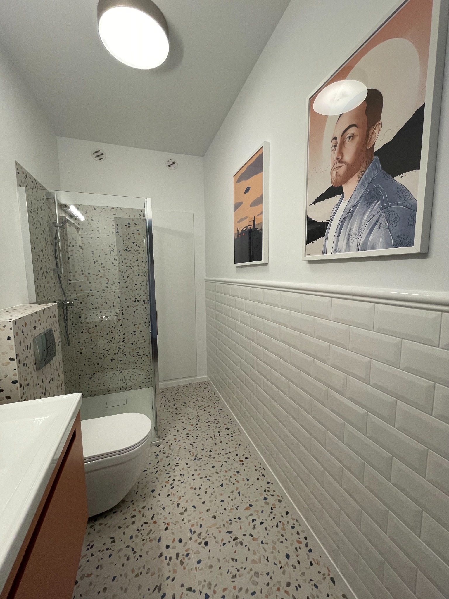 Nowoczesna łazienka z kabiną prysznicową, umywalką z szafką w kolorze terakoty, toaletą i terakotową podłogą. Na ścianach białe płytki typu metro i dwa obrazy w białych ramach.