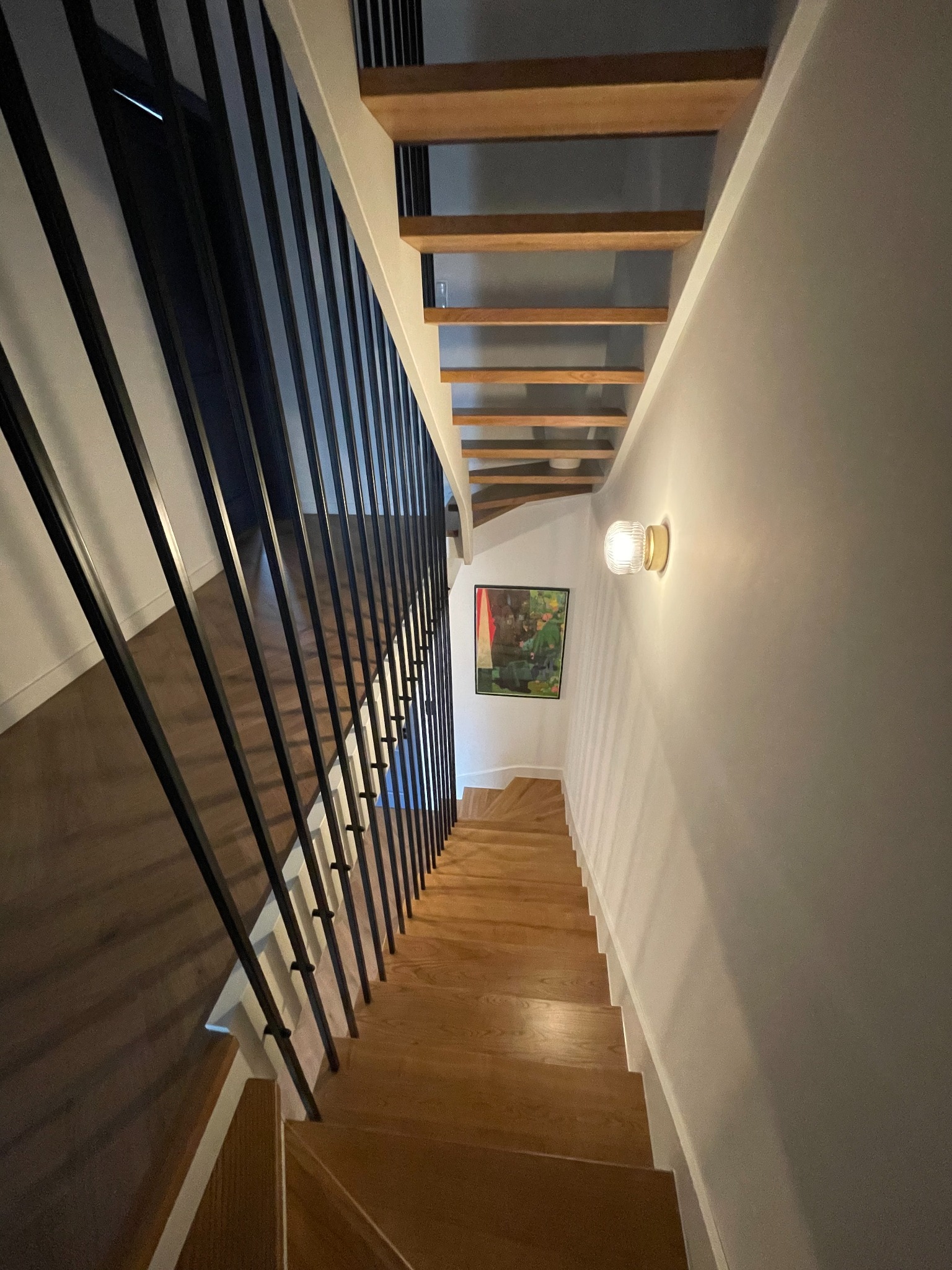 Drewniane schody z czarną metalową balustradą i białą ścianą, oświetlone kinkietem z fakturą, z obrazem na ścianie w tle.