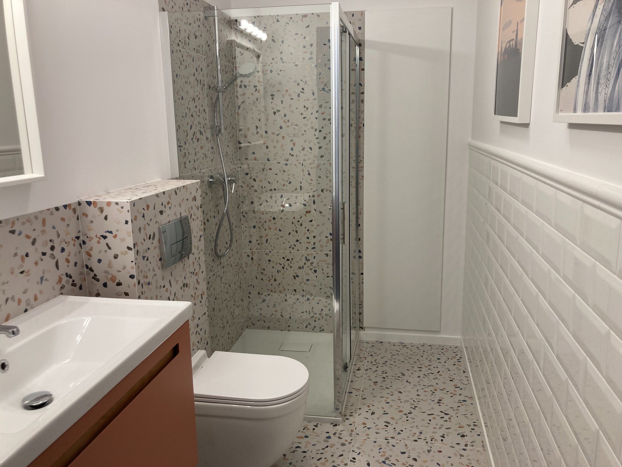 Nowoczesna łazienka z kabiną prysznicową, umywalką na szafce w kolorze terakoty i toaletą, wykończona płytkami lastryko na podłodze i części ścian, oraz białymi płytkami imitującymi cegłę na dolnej...