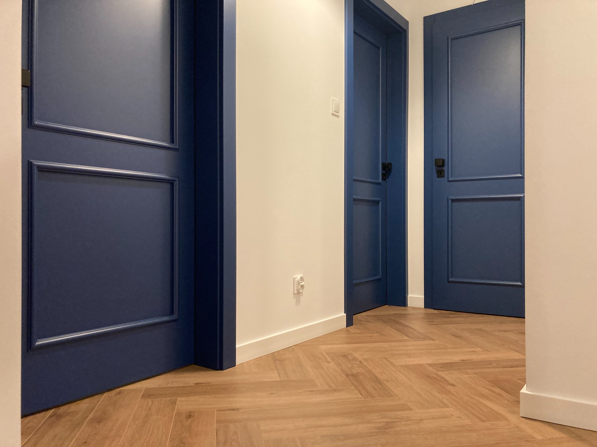 Korytarz z trzema parami ciemnoniebieskich drzwi z dekoracyjnymi listwami, ułożona w jodełkę podłoga z paneli w odcieniach brązu, białe ściany i listwy przypodłogowe, widoczne gniazdko elektryczne.