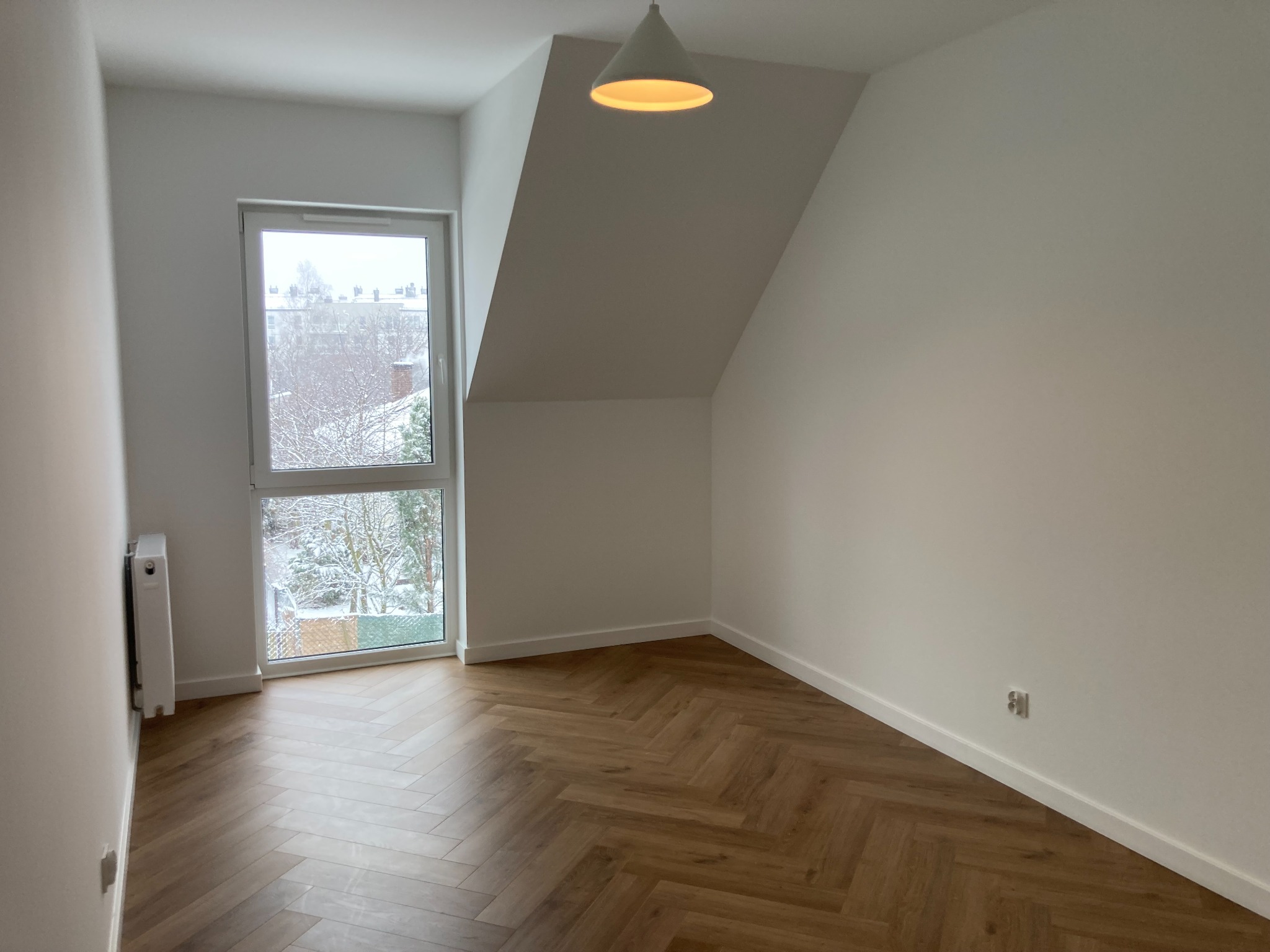 Pusty pokój na poddaszu po remoncie: parkiet ułożony w jodełkę, białe ściany, okno z widokiem na zimowy krajobraz, minimalistyczna lampa wisząca.