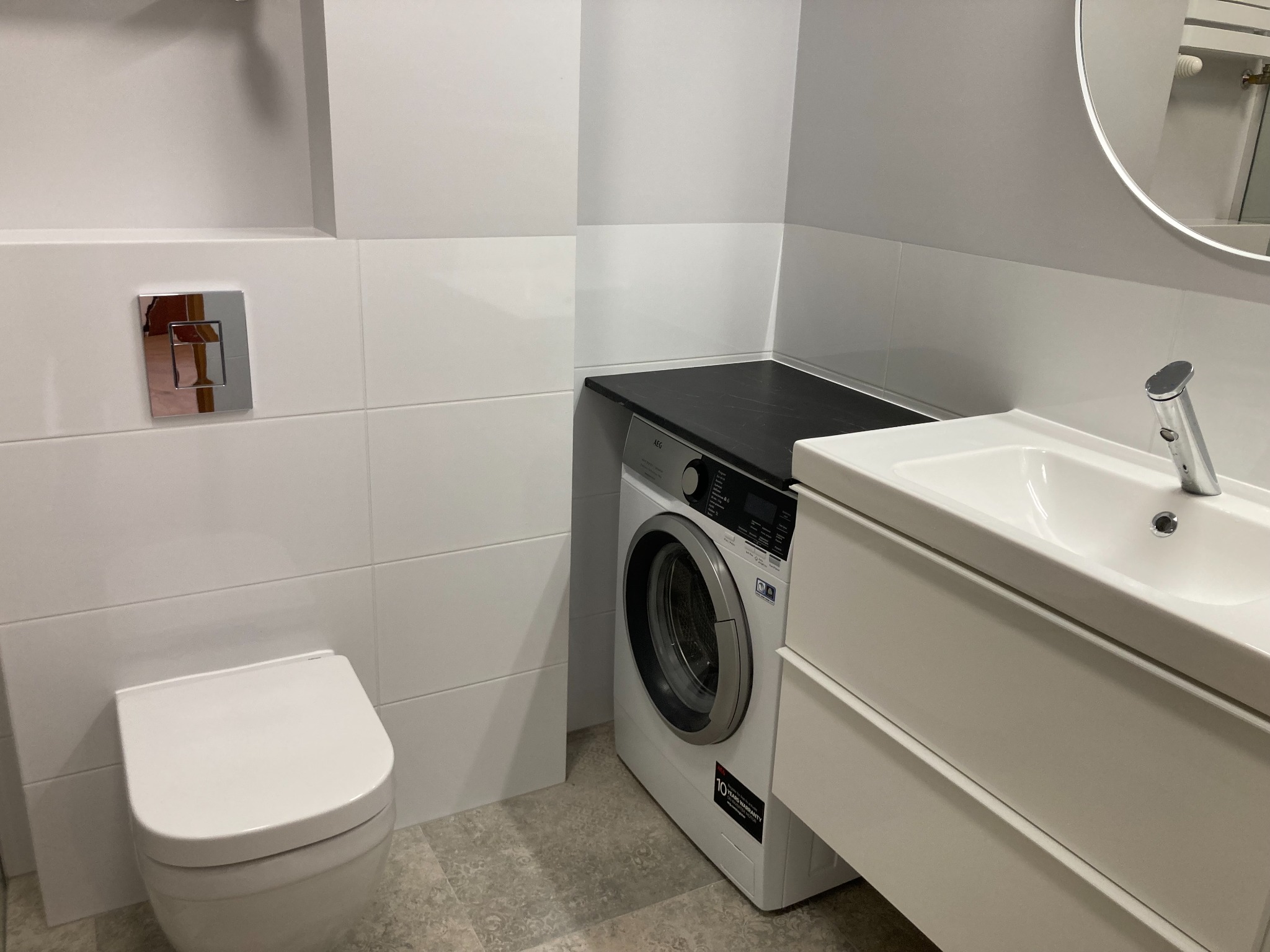 Nowoczesna łazienka z białymi płytkami, podwieszaną toaletą, pralką pod blatem i umywalką z szafką, wykończona w minimalistycznym stylu.