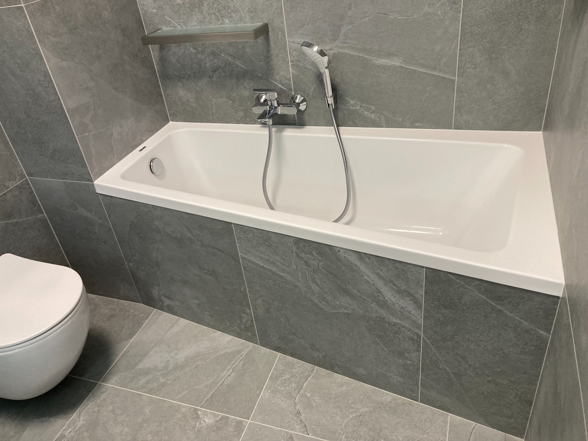 Nowoczesna łazienka z szarą, kamienną okładziną, białą wanną z chromowaną baterią i deszczownicą, oraz wiszącą półką i toaletą, oświetlenie górne.