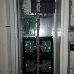 Szafa sterownicza z modułami elektroniki, przewodami i zasilaczem, widoczne płytki drukowane i połączenia kablowe.