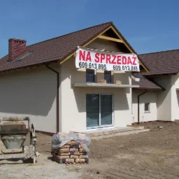 Nowo wybudowany dom z brązowym dachem i beżową elewacją, z banerem 'Na sprzedaż' nad oknem, widoczna betoniarka i worki cementu na placu budowy.