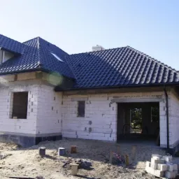 Niedokończony dom jednorodzinny z białych bloczków, z ciemną dachówką i otwartym garażem, widoczne puszki po farbie i paleta bloczków na piaszczystym terenie budowy.