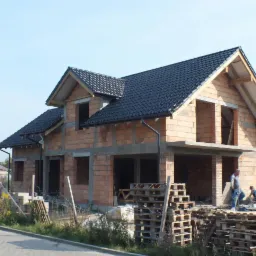 Wznoszenie dwukondygnacyjnego domu z czerwonej cegły, z czarnym dachem z dachówki, widoczne palety z materiałami budowlanymi i pracujący robotnicy.