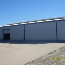 Gotowa hala magazynowa z szarej blachy trapezowej, z częściowo otwartą bramą rolowaną, widoczna data wykonania zdjęcia