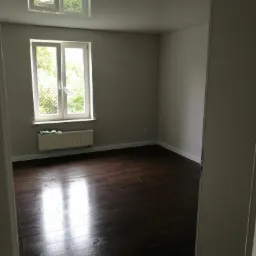 Pokój po odnowieniu z ciemną podłogą z paneli imitujących drewno, jasnymi ścianami i oknem z widokiem na zieleń, widziany zza futryny.