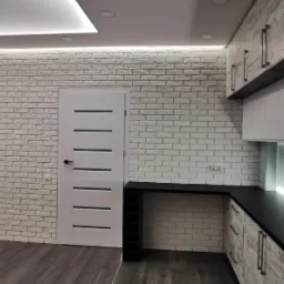 Nowoczesna kuchnia z białymi ceglanymi ścianami, szarymi panelami podłogowymi, białymi szafkami z szarymi frontami i czarnym blatem, oświetleniem LED pod szafkami i na suficie.