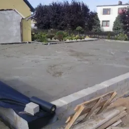 Wylana płyta fundamentowa z izolacją i obmurowaniem z bloczków betonowych. Widoczne deski szalunkowe. W tle budynki mieszkalne i zieleń.