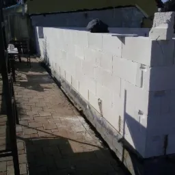 Wznoszenie ściany z białych bloczków, widoczne detale łączeń. Częściowo zadaszony obszar budowy z kostką brukową i elementami drewnianymi w tle.