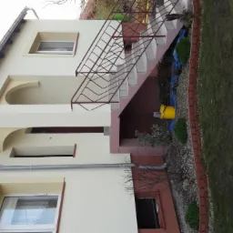 Jasna elewacja domu jednorodzinnego z dekoracyjnymi łukami nad oknami i wejściem, widoczne schody zewnętrzne z brązową balustradą oraz żółte wiadro przy ścianie.