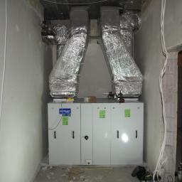 Cool System Systemy Wentylacji i Klimatyzacji - Biała centrala wentylacyjna Komfovent z dwoma srebrnymi przewodami wentylacyjnymi w surowym pomieszczeniu z przewodami na ścianie. Widoczne narzędzia i elementy montażowe.