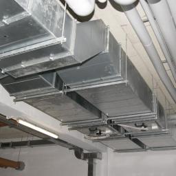 Cool System Systemy Wentylacji i Klimatyzacji - Sufit z widocznymi, zainstalowanymi kanałami wentylacyjnymi o przekroju prostokątnym, rurami oraz elementami instalacji elektrycznej. Widok z dołu.