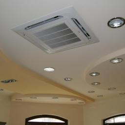 Cool System Systemy Wentylacji i Klimatyzacji - Kasetonowa klimatyzacja w suficie podwieszanym z oświetleniem punktowym. Widok na sufit z zamontowanym urządzeniem w jasnym wnętrzu.