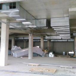 Cool System Systemy Wentylacji i Klimatyzacji - Montaż kanałów wentylacyjnych pod stropem budynku komercyjnego. Widoczne elementy konstrukcyjne, częściowo rozpakowane kanały i pracownik w tle.