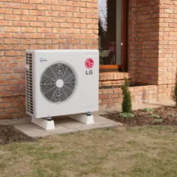 Biała jednostka zewnętrzna klimatyzacji LG Inverter, zamontowana przy ceglanym budynku z oknem. Widoczne krzewy i trawnik.