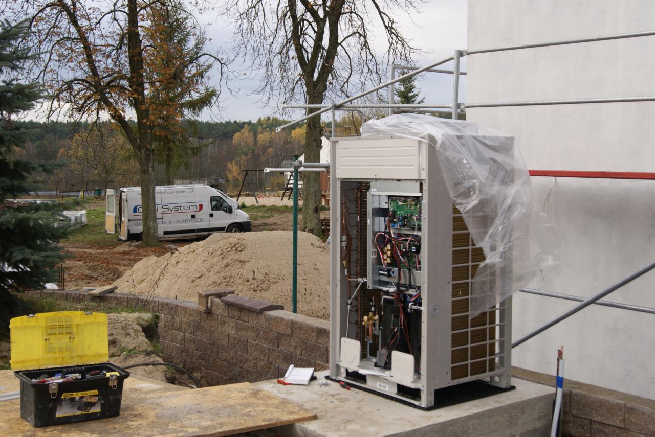 Otwarta obudowa klimatyzatora zewnętrznego na fundamencie, narzędzia w kufrze, auto firmowe w tle. Widoczna instalacja wewnątrz urządzenia.