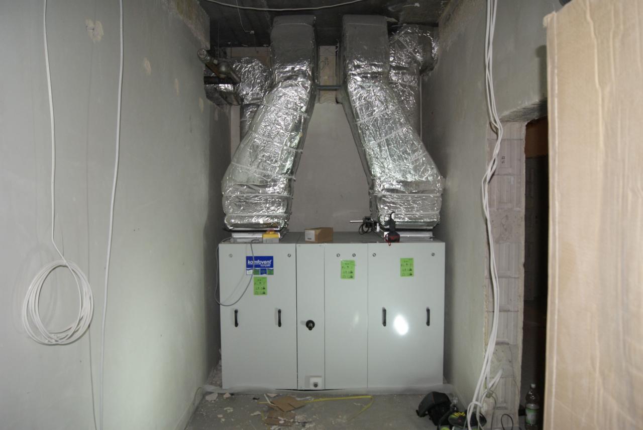 Biała centrala wentylacyjna Komfovent z dwoma srebrnymi przewodami wentylacyjnymi w surowym pomieszczeniu z przewodami na ścianie. Widoczne narzędzia i elementy montażowe.