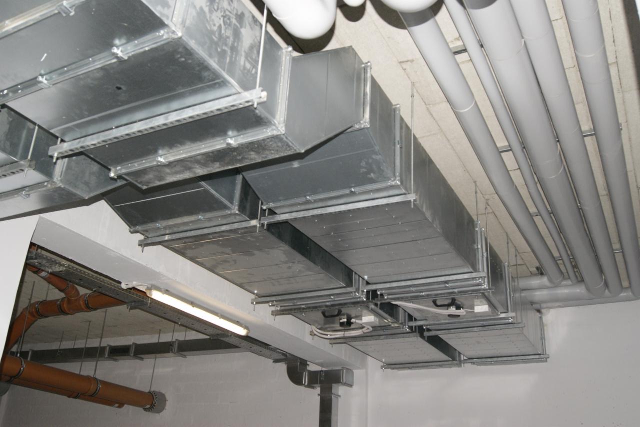 Sufit z widocznymi, zainstalowanymi kanałami wentylacyjnymi o przekroju prostokątnym, rurami oraz elementami instalacji elektrycznej. Widok z dołu.