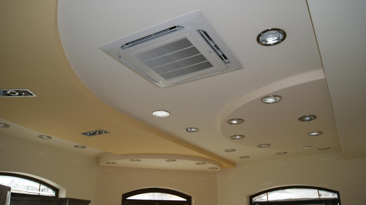 Kasetonowa klimatyzacja w suficie podwieszanym z oświetleniem punktowym. Widok na sufit z zamontowanym urządzeniem w jasnym wnętrzu.