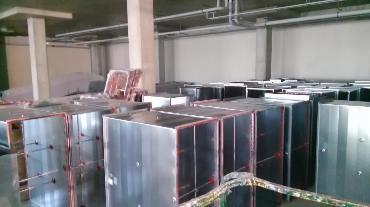 Magazyn z dużą ilością metalowych obudów wentylacyjnych, prawdopodobnie central wentylacyjnych, ułożonych w rzędach. Widoczne rury pod sufitem i słupy konstrukcyjne.