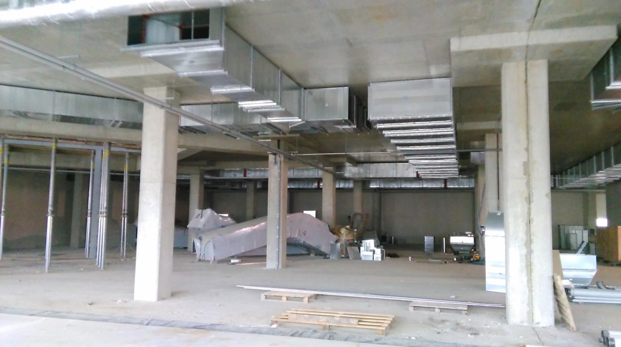 Montaż kanałów wentylacyjnych pod stropem budynku komercyjnego. Widoczne elementy konstrukcyjne, częściowo rozpakowane kanały i pracownik w tle.