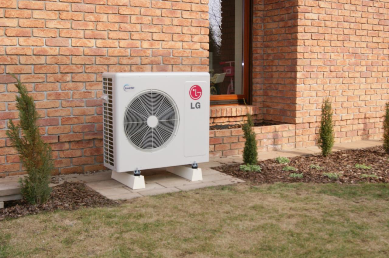 Biała jednostka zewnętrzna klimatyzacji LG Inverter, zamontowana przy ceglanym budynku z oknem. Widoczne krzewy i trawnik.
