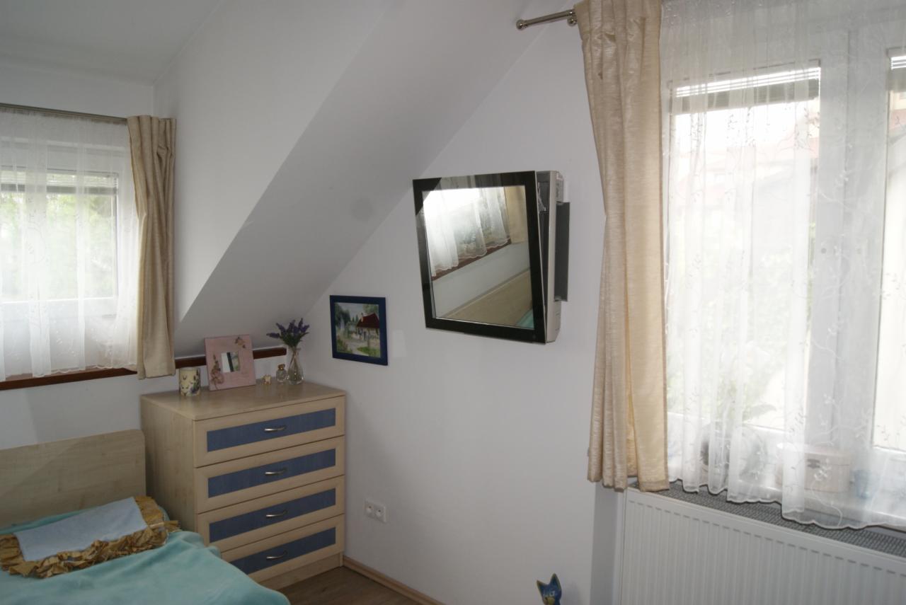 Klimatyzator ścienny w pokoju na poddaszu, zamontowany obok okna z firanką i zasłoną. Widoczna komoda, obrazek i dekoracje. Ujęcie z dołu.