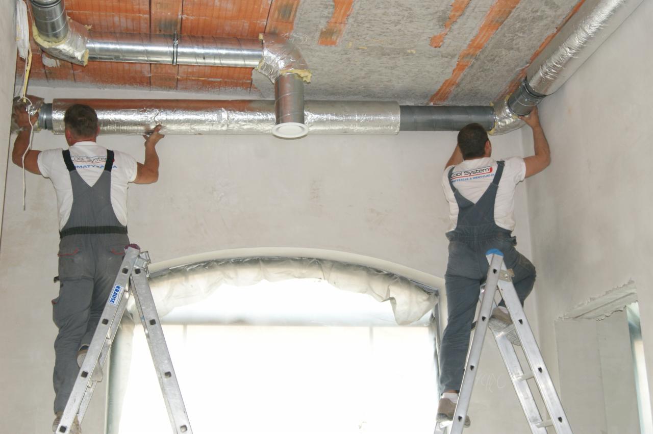 Dwóch monterów na drabinach instaluje srebrne rury wentylacyjne pod ceglanym stropem w jasnym, surowym wnętrzu. Widoczne łączenia rur i izolacja.