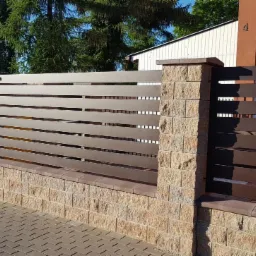 Nowoczesne, poziome przęsła w kolorze brązowym, osadzone na słupkach z kamienia łupanego, tworzące ogrodzenie posesji z kostki brukowej.