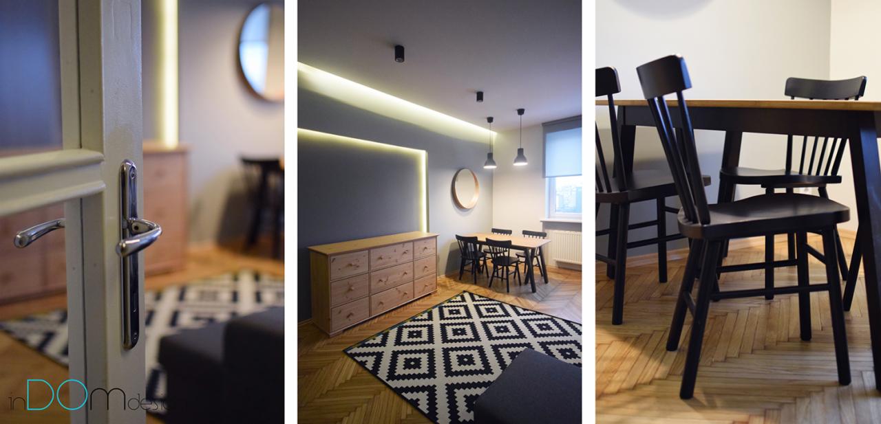 Wnętrze jadalni z drewnianą komodą, czarnym dywanem w geometryczne wzory, stołem i krzesłami, oświetleniem LED oraz okrągłym lustrem, widziane przez uchylone drzwi.