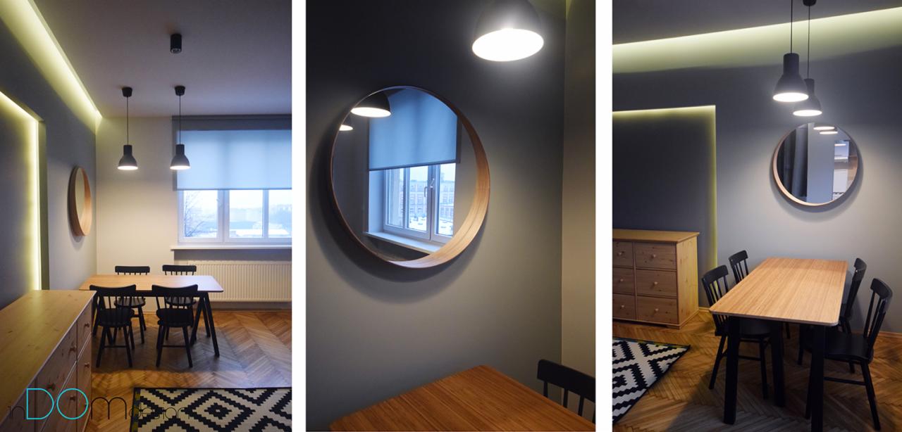 Jadalnia z drewnianym stołem i czarnymi krzesłami, okrągłe lustro na ścianie, parkiet w jodełkę i czarno-biały dywan w geometryczne wzory, oświetlenie punktowe i listwa LED.