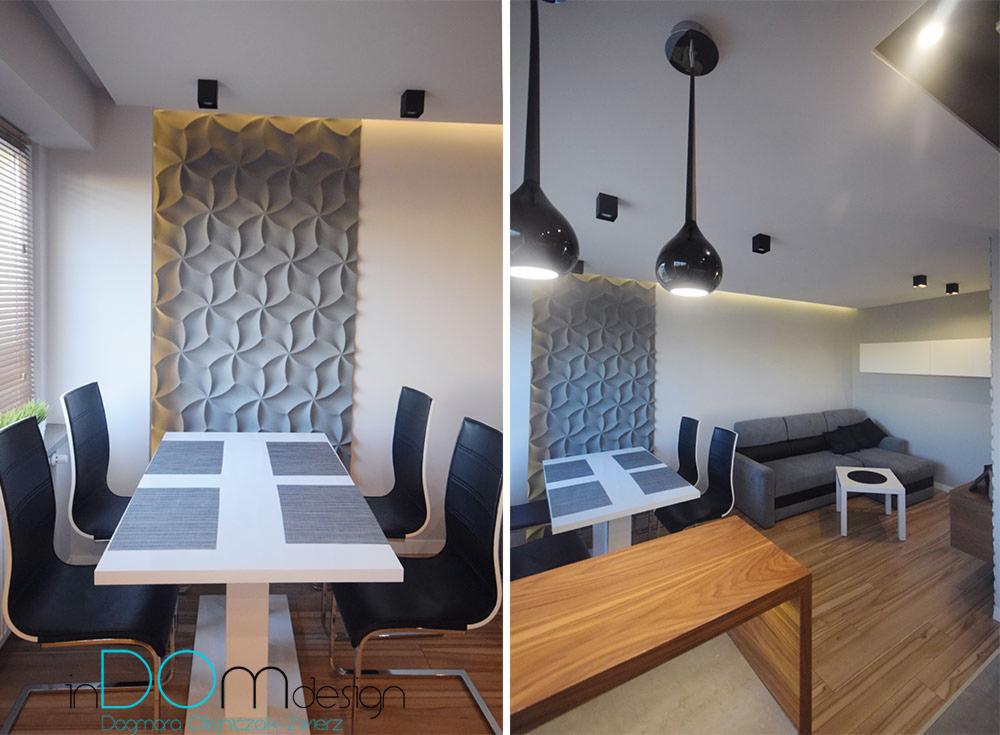 Nowoczesna jadalnia z białym stołem, czarnymi krzesłami i dekoracyjną trójwymiarową ścianą z geometrycznym wzorem, oświetlona wiszącymi lampami w kształcie kropli.