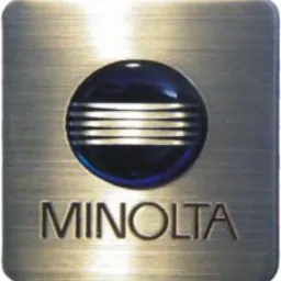 Metalowa plakietka z logo Minolta, okrągły element z niebieskim tłem i poziomymi srebrnymi paskami, napis MINOLTA wytłoczony w metalu.