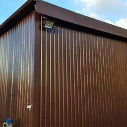 Brązowa hala z płyt warstwowych pod błękitnym niebem, widoczny fragment rynny i lampa oświetleniowa. Charakterystyczne pionowe przetłoczenia blachy.