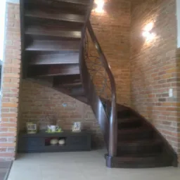 Gięte, drewniane schody z ozdobną, metalową balustradą, umieszczone we wnętrzu z ceglanymi ścianami i jasną podłogą.