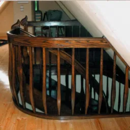 Gięta balustrada z ciemnego drewna, z dekoracyjnymi zakończeniami słupków, na drewnianych schodach w poddaszowym pomieszczeniu.