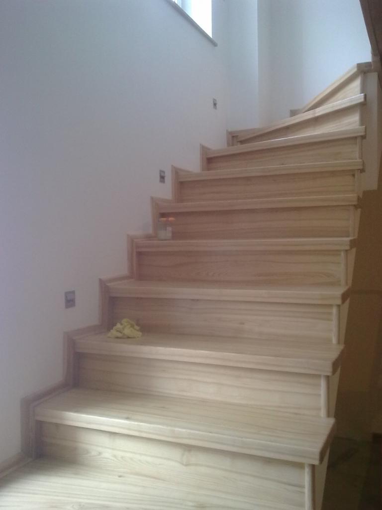 Jasne, niedokończone drewniane schody na betonowej konstrukcji, widoczne ślady prac wykończeniowych w postaci plam i ścierek na stopniach.