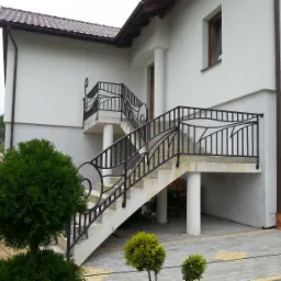 Nowoczesne, metalowe balustrady schodowe z dekoracyjnymi elementami, zamontowane przy wejściu do domu z jasną elewacją i brązowymi oknami.