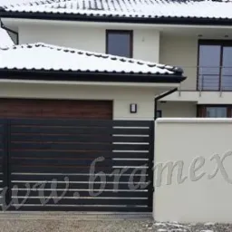 Nowoczesna, pozioma brama wjazdowa i furtka z czarnych paneli na tle domu z białymi ścianami i śniegiem na dachu. Widoczny adres strony internetowej.