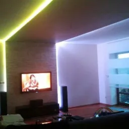 Salon z podwieszanym sufitem i oświetleniem LED wzdłuż krawędzi, telewizor na ścianie z dekoracyjnej cegły, duże okno z roletami.