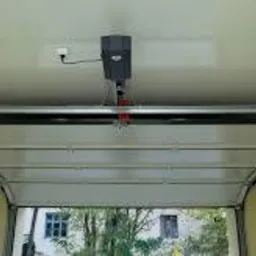 Wnętrze garażu z zamontowaną automatyką do bramy segmentowej, widoczny napęd centralny, szyna prowadząca i oświetlenie boczne. Brama w pozycji otwartej, z widokiem na zewnątrz.