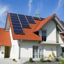 Dom jednorodzinny z panelami słonecznymi na dachu z czerwonej dachówki, widok z poziomu ogrodu w słoneczny dzień, błękitne niebo z lekkimi chmurami w tle.