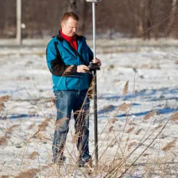 Geodeta w niebieskiej kurtce, obsługujący precyzyjny odbiornik GNSS na zasnieżonym polu z uschniętymi trawami.
