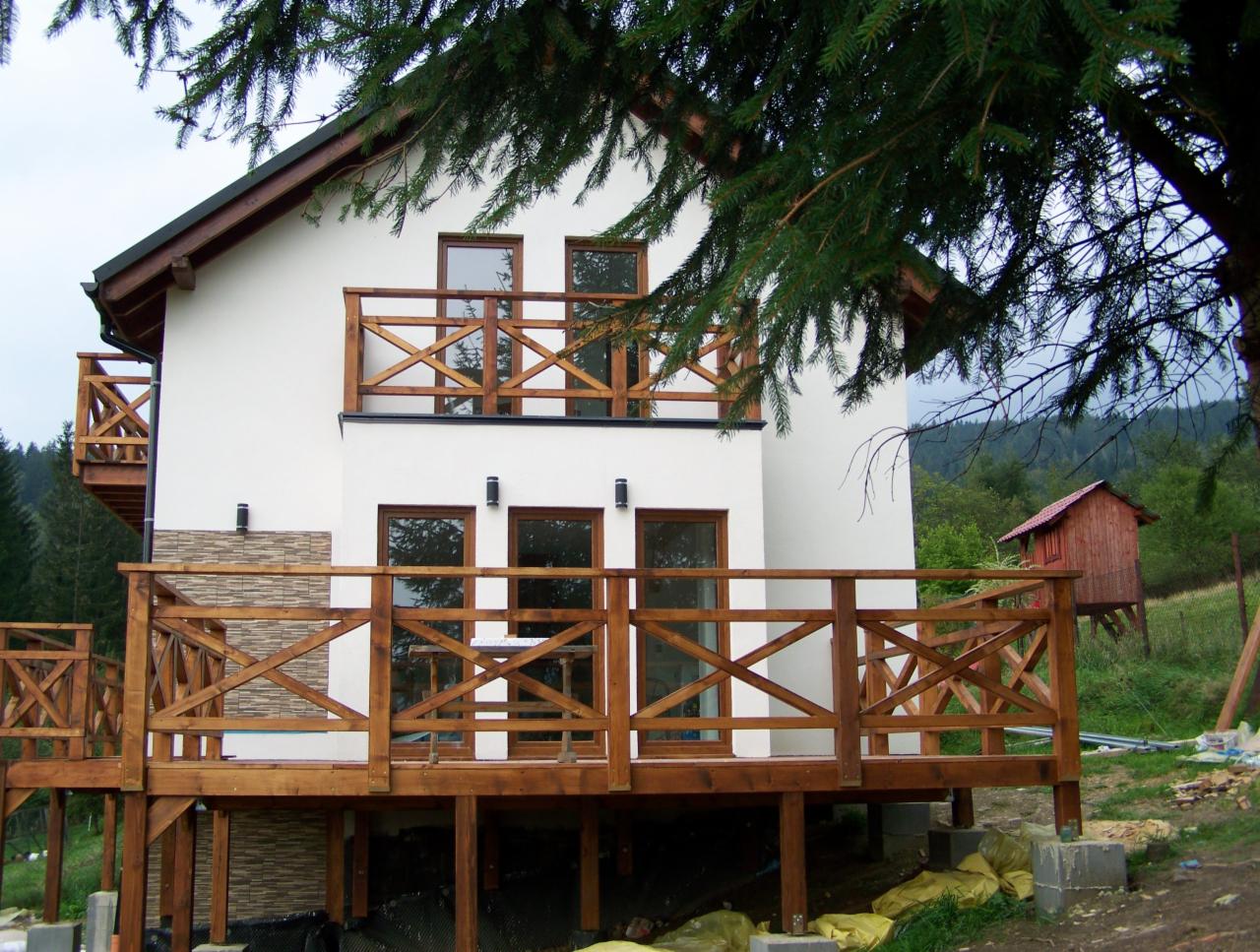 Nowoczesny dom z jasną elewacją i drewnianymi balustradami balkonów, wkomponowany w górski krajobraz z widocznym fragmentem lasu i małą budowlą w tle.