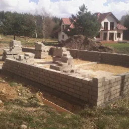 Budowa domu: wznoszenie ścian fundamentowych z bloczków betonowych. W tle dom z czerwonym dachem i drzewa. Widoczny wykop pod fundamenty.