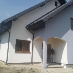 Dwukondygnacyjny dom jednorodzinny z szarą dachówką i białymi ścianami po lewej stronie oraz szarymi po prawej, pracownik maluje ściany przy wejściu, widoczne puszki z farbą.