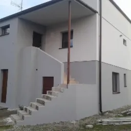 Dwukondygnacyjny dom po remoncie elewacji, z widocznymi schodami zewnętrznymi i świeżo położonym tynkiem w odcieniach szarości i bieli, rury spustowe w kolorze brązowym.